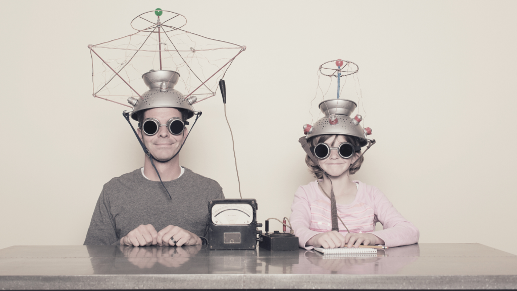 un père et une fille assis de face à table semblent être en train de réaliser une expérience scientifique loufoque à base de chapeaux magnétiques faits maison et d'un multimètre.